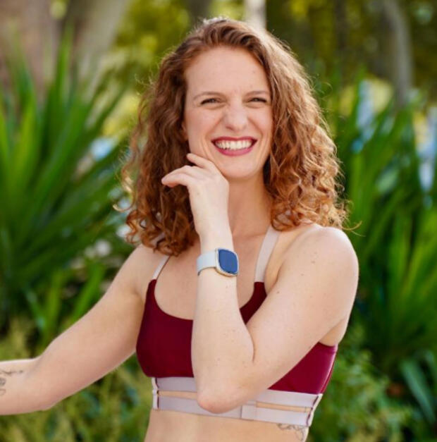 bio picture of Camila smiling, wearing a red top on a leafy background.